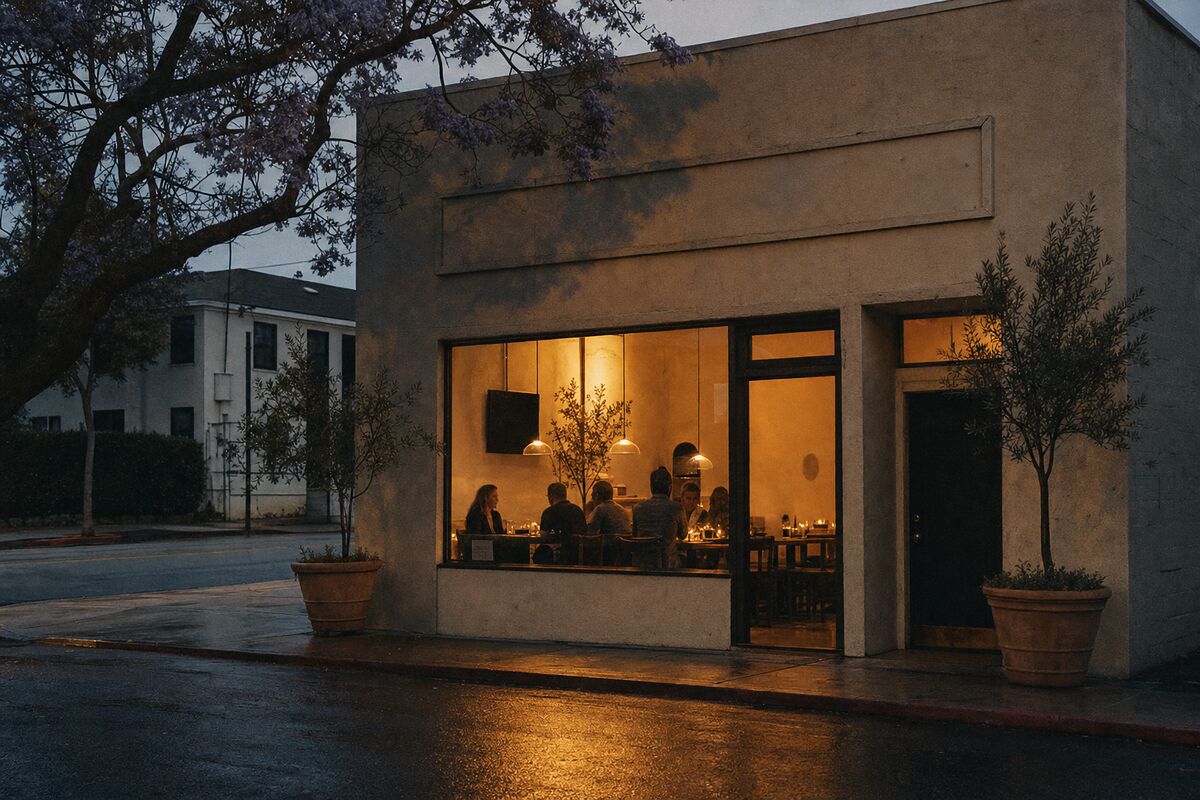 A small independent restaurant on a California street at dusk, its front window glowing warm amber with diners inside.