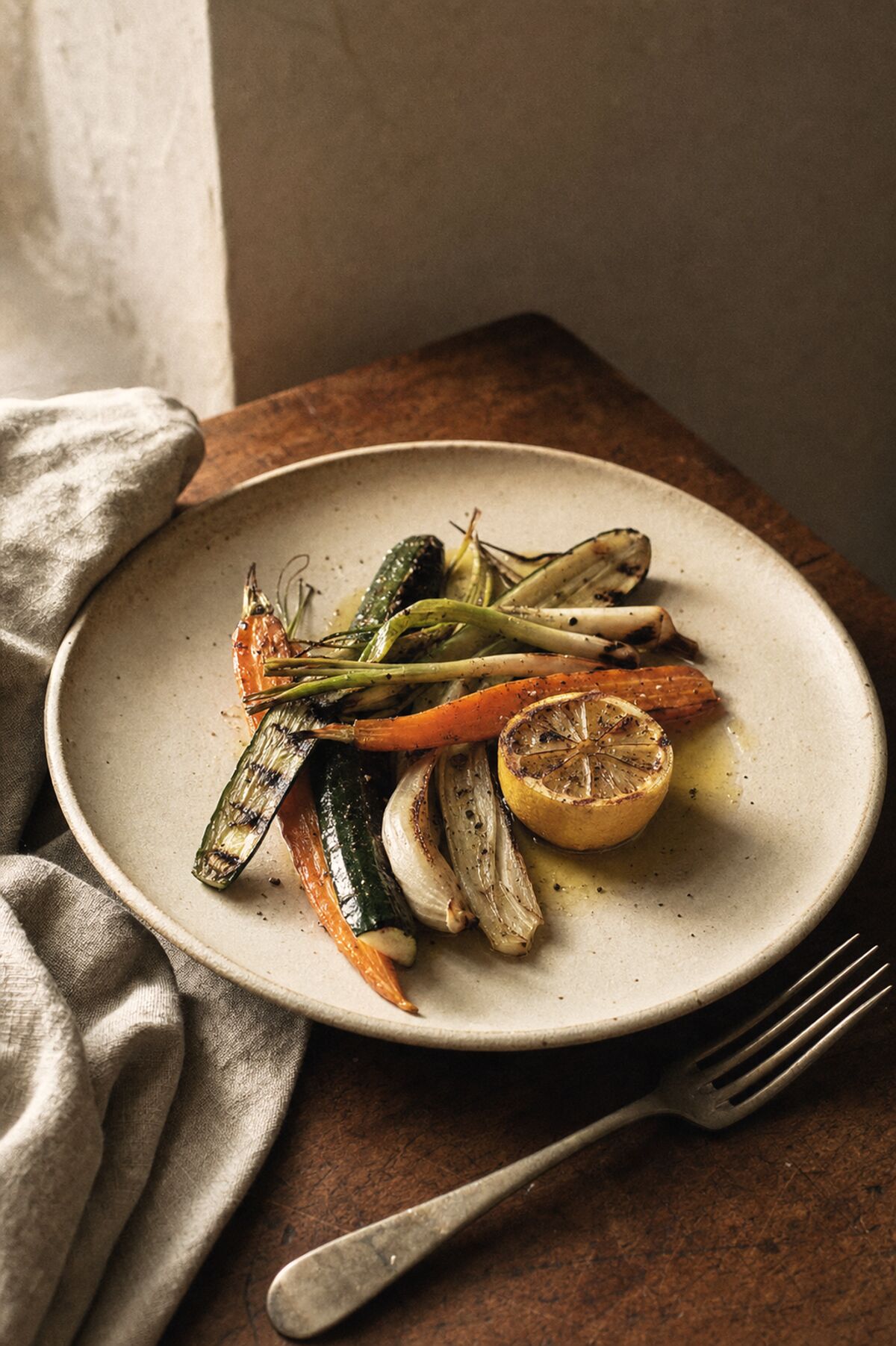 A plate of grilled market vegetables with charred lemon and olive oil on a matte ceramic plate, on a worn walnut table.